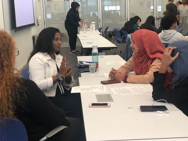A group of women sitting a table with papers in front of them. They're talking to each other, and other women doing the same are in the background. They are indoors, a glass wall revealing a hallway is behind them.